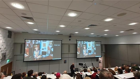 A lecture hall, photo taken from the back of the room. Two huge projector screens on the wall