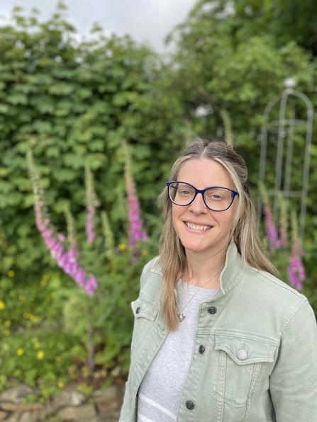 A lady in a green jacket with glasses on stands in front of a hedgerow filled with flowering fox gloves