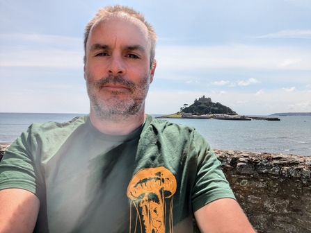 A man in the green tshirt with a graphic of an orange jellyfish on it, stood by a wall in front of the sea, with a small island in the background