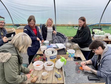 People sat & stood around a big wooden table making food in a polytunnel