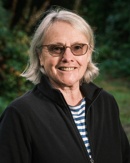 A woman with short, grey hair wearing glasses, a black jacket, and a striped shirt, smiles at the camera. The background is outdoors, with greenery out of focus.