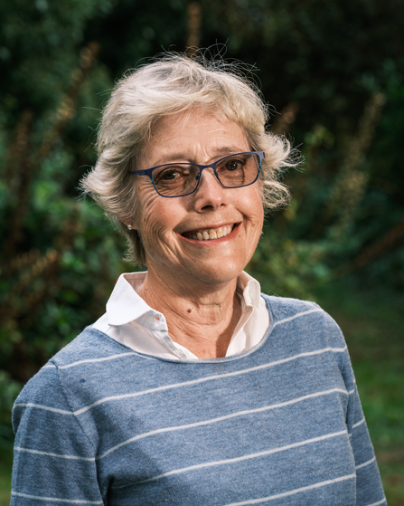 A woman with short grey hair, wearing glasses and a white shirt with a blue striped jumper over the top, smiles at the camera.  The background is outdoors, with greenery out of focus.