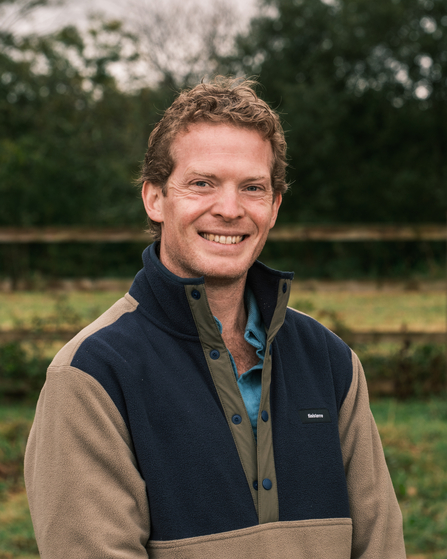 A man with short, curly hair smiles at the camera, wearing a tan and navy fleece jacket. The background is outdoors with trees and fencing in soft focus.