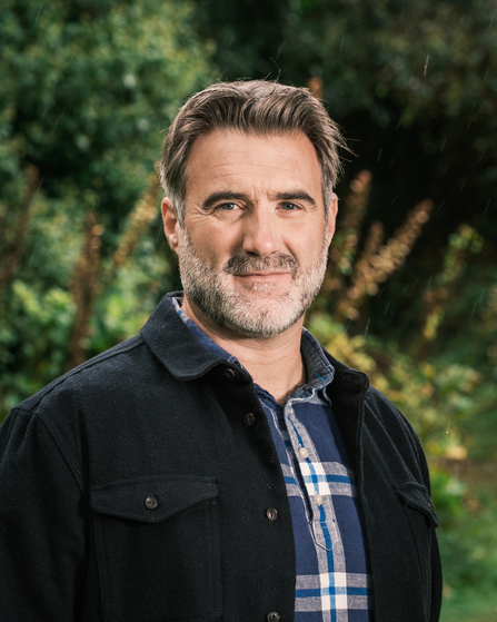 A man with short grey hair and a light beard wearing a black jacket over a plaid shirt smiles and looks at the camera. The background is outdoors, with the greenery out of focus.