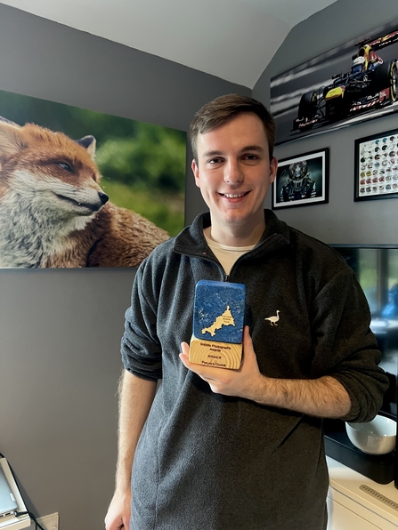 A brown-haired man smiling while holding a "People's Choice Winner" award from Cornwall Wildlife Trust's Wildlife Photography Awards, standing in a room with wildlife and motorsport-themed artwork on the walls.