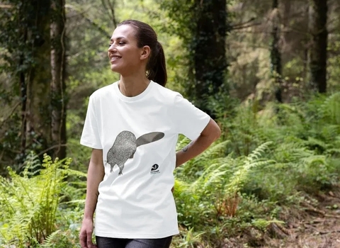 A woman in a woods wearing a white tshirt with an illustration of a beaver on it